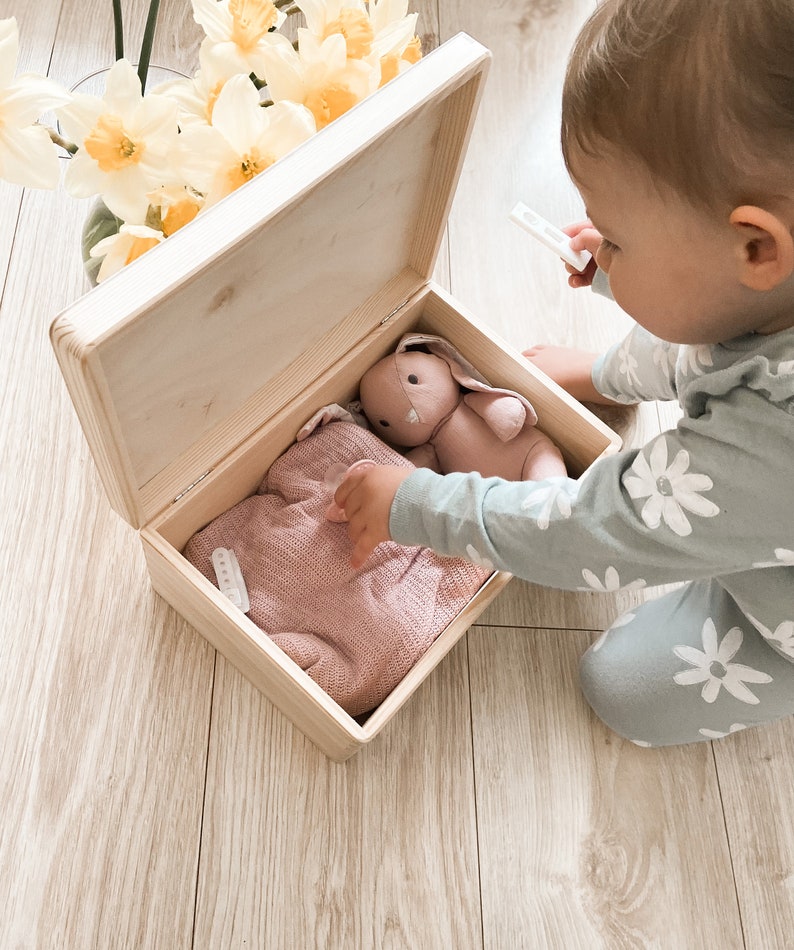 Peut inclure: Une bo&icirc;te en bois avec un pull en tricot rose clair et un jouet de lapin rose &agrave; l'int&eacute;rieur. La bo&icirc;te est ouverte et un enfant met la main dans la bo&icirc;te.