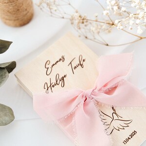 K&ouml;nnte beinhalten: Eine Holzkiste mit einem rosa Band und einem Taubenmotiv. Die Kiste ist mit dem Text "Emma's Heilige Taufe" und dem Datum "13.04.2025" graviert.