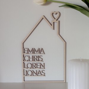 May include: A wooden house-shaped sign with the names Emma, Chris, Loren, and Jonas written on it. The sign has a heart-shaped cutout on top of the roof.