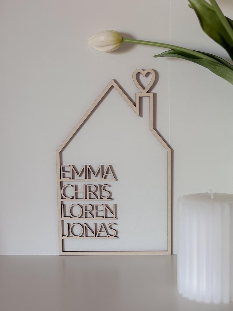 May include: A wooden house-shaped sign with the names Emma, Chris, Loren, and Jonas written on it. The sign has a heart-shaped cutout on top of the roof.