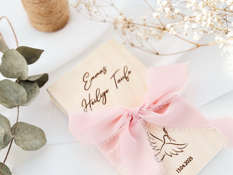 May include: A wooden box with a pink ribbon and a dove design. The box is engraved with the text "Emma's Heilige Taufe" and the date "13.04.2025".
