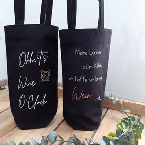 Könnte beinhalten: Zwei schwarze Canvas-Weinflaschentaschen. Die Tasche links hat weißen Text, der "Oh, it's Wine O'Clock" mit einer goldenen Uhrgrafik sagt. Die Tasche rechts hat weißen Text, der "Meine Laune ist im Keller, ich hoffe sie bringt Wein mit" sagt.