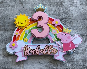 May include: A pink and gold cake topper featuring a rainbow, a sun, a number 3, a crown, a pink pig with wings and a wand, and the name "Isabella".