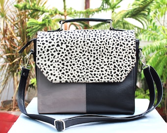 May include: A black and gray leather satchel with a white and black spotted flap. The bag has a top handle and a long adjustable shoulder strap.