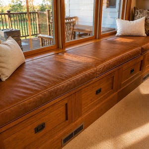 May include: A built-in wooden window seat featuring a smooth brown leather cushion and integrated storage drawers with dark metal pulls. The seat is accented with several decorative throw pillows in cream and patterned brown fabric.