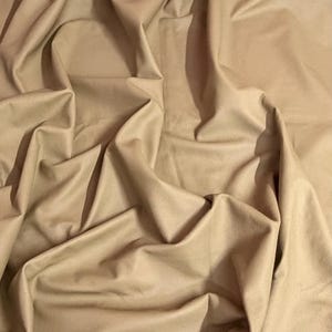 Può includere: Un grande pezzo di pelle color marrone chiaro con una finitura liscia e opaca. Il materiale è drappeggiato in pieghe profonde e fluide che creano una serie di ombre e riflessi sulla superficie, enfatizzando la consistenza morbida della pelle.