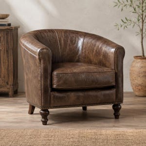 May include: A brown leather barrel chair with a curved backrest, a thick seat cushion, and dark wooden turned legs. The upholstery features a distressed finish with visible grain texture and a row of small metal nailhead trim along the base.