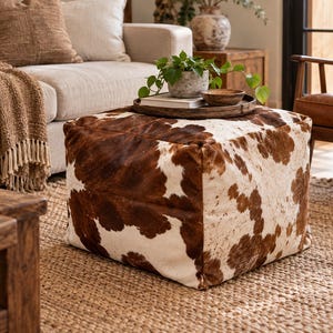 May include: A square ottoman upholstered in a cowhide pattern featuring irregular brown and white patches. The piece has a structured, boxy shape with visible seams along the edges, showcasing a natural hair-on-hide texture throughout the surface.