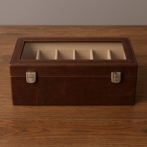 Può includere: Una scatola per orologi in pelle marrone con un coperchio in vetro trasparente e cinque scomparti. La scatola ha una forma rettangolare con chiusure color argento e un interno beige chiaro. La scatola è progettata per riporre e proteggere gli orologi.