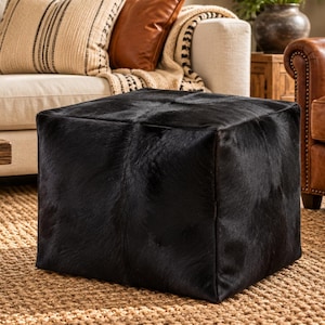May include: A square ottoman upholstered in black hair-on-hide leather. The cube-shaped piece features a smooth, dark, textured surface with visible natural hair patterns, providing a solid and structured appearance for the living space.