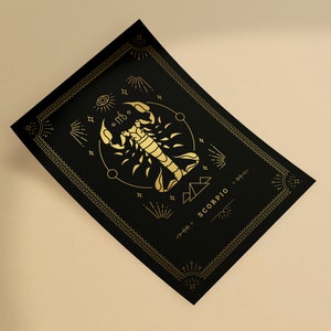 May include: A black rectangular card with a gold scorpion zodiac design. The design includes the word "SCORPIO" and other celestial symbols. The card has a decorative gold border and is angled on a beige surface.
