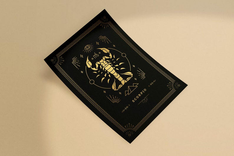 May include: A black rectangular card with a gold scorpion zodiac design. The design includes the word "SCORPIO" and other celestial symbols. The card has a decorative gold border and is angled on a beige surface.