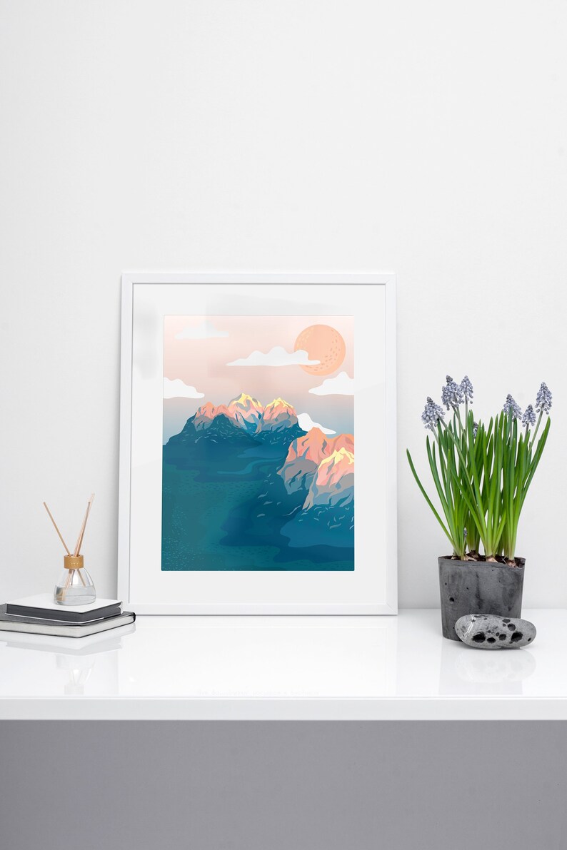 May include: A framed art print featuring a mountain landscape with a peach-colored sun and clouds. The mountains are blue and peach, set against a light blue sky. A potted plant with purple flowers sits to the right.