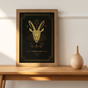 May include: A framed art print featuring a gold Capricorn zodiac symbol on a black background. The print is in a light brown wooden frame and is displayed on a wooden table. A small vase and sphere are also on the table.