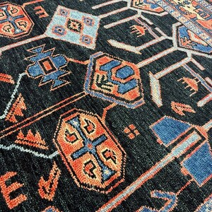 May include: Close-up of a dark blue rug with a geometric pattern. The rug features repeating shapes in shades of orange, blue, and light blue. The design includes diamond, cross, and rectangular shapes. The rug has a textured surface.