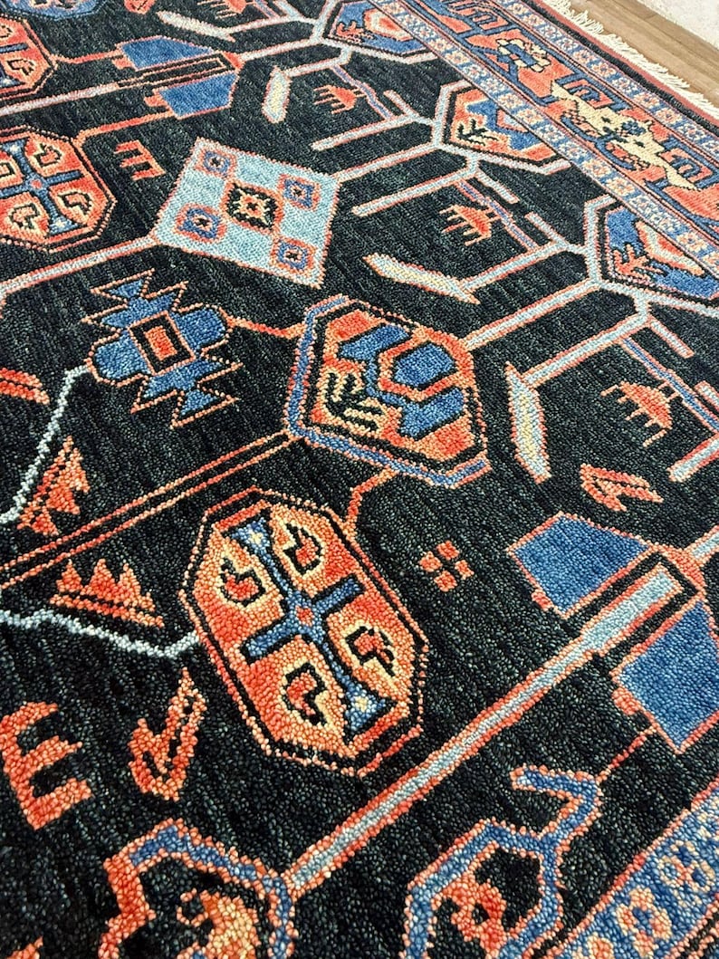 May include: Close-up of a dark blue rug with a geometric pattern. The rug features repeating shapes in shades of orange, blue, and light blue. The design includes diamond, cross, and rectangular shapes. The rug has a textured surface.