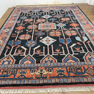 May include: A large, rectangular rug with a dark background and intricate geometric patterns in shades of orange, blue, and white. The rug has a detailed border and fringe along the edges, suitable for home decor.