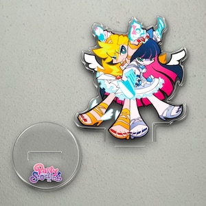 Puede incluir: Un soporte de acrílico transparente con una colorida ilustración de estilo anime de dos personajes con alas de ángel. Uno tiene el pelo amarillo y el otro el pelo azul oscuro. El soporte incluye una base separada con el texto "Panty & Stocking".