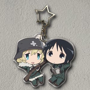 Puede incluir: Un llavero acrílico transparente con dos personajes de estilo anime. Uno tiene cabello rubio, un sombrero militar con una cruz y un rifle. El otro tiene cabello negro y está leyendo un libro. El llavero tiene un cierre en forma de estrella.