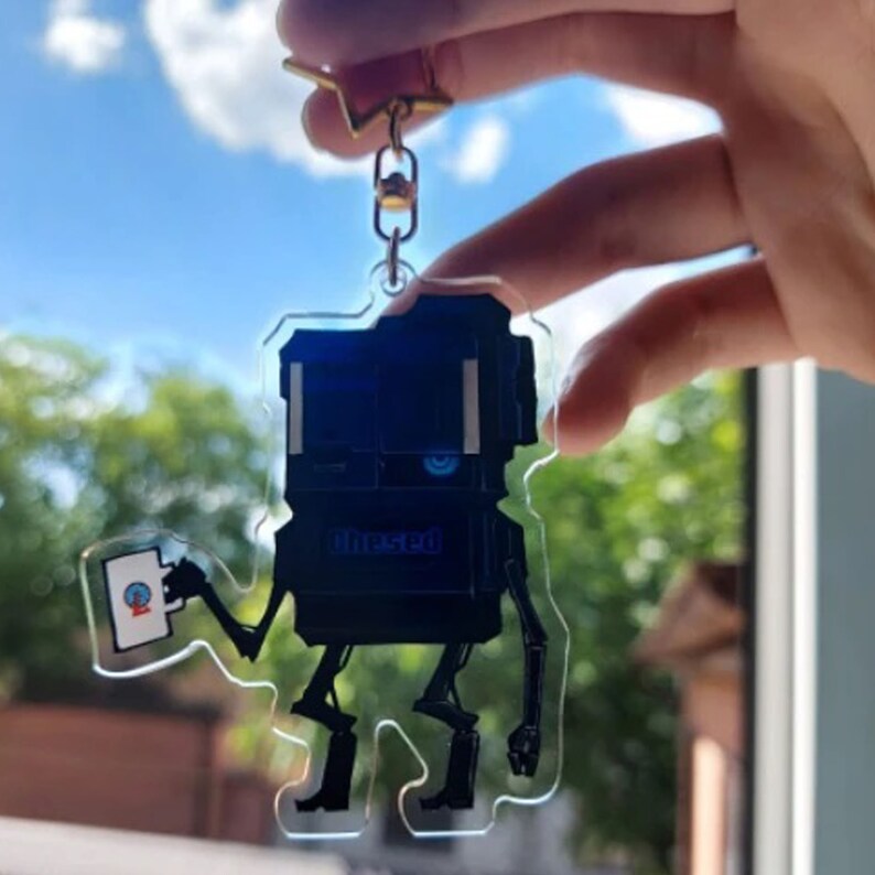 May include: A clear acrylic keyring featuring a robot-like figure with a dark blue body and black limbs. The keyring has a gold-coloured star-shaped clasp and a small card in its hand. The word "thesed" is printed on the robot's body.