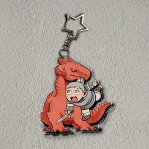 Op de afbeelding: Een heldere acryl sleutelhanger met een cartoon illustratie van een rode draak en een kleine ridder met een zwaard. De sleutelhanger heeft een zilveren stervormige sluiting.