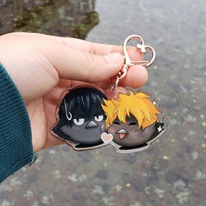 May include: A clear acrylic keychain with two cartoon bird characters. One bird has black hair and a grumpy expression, the other has orange hair and a happy expression. The keychain has a rose gold heart-shaped clasp.