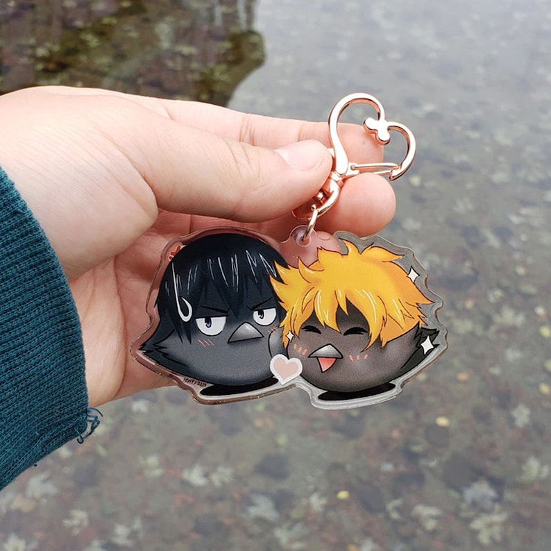 May include: A clear acrylic keychain with two cartoon bird characters. One bird has black hair and a grumpy expression, the other has orange hair and a happy expression. The keychain has a rose gold heart-shaped clasp.