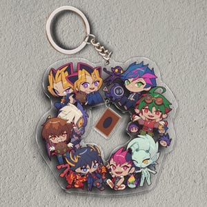 May include: A clear acrylic keychain featuring a collection of cartoon characters in a circular arrangement. The characters have large eyes and colorful outfits. A small card is in the center. The keychain has a silver chain and ring.