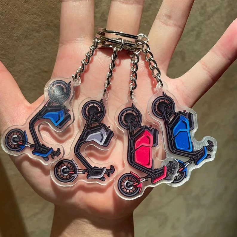 May include: Four clear acrylic scooter keyrings with silver keyrings and chains. Each keyring features a scooter design in different colours: blue, grey, red, and blue. The scooters have black wheels and frames.