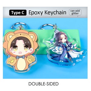 May include: Two double-sided epoxy keychains. One features a cartoon character in a bear costume, the other a character in blue robes. The keychains have silver key rings and the text "Type C Epoxy Keychain" and "can add glitter". The word "DOUBLE-SIDED" is also visible.