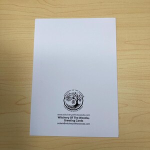 May include: A white greeting card with the text "Witchery Of The Woods&reg; Greeting Cards" and a circular logo featuring a tree and other symbols. The card is on a light brown wooden surface.