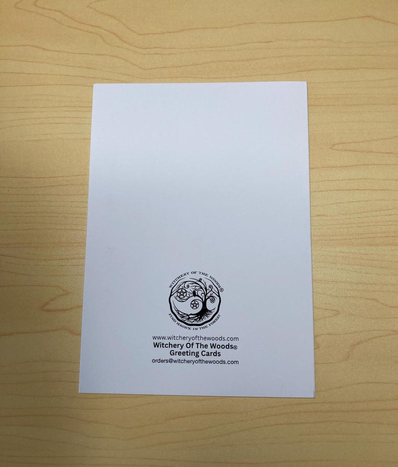 May include: A white greeting card with the text "Witchery Of The Woods&reg; Greeting Cards" and a circular logo featuring a tree and other symbols. The card is on a light brown wooden surface.