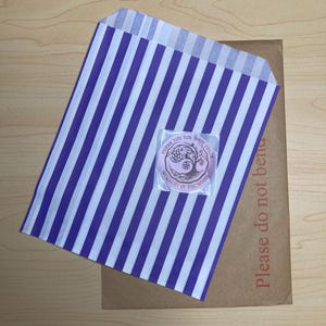 May include: A purple and white striped paper bag with a small sticker on top. The sticker has text that reads "Thank you for your order" and "Witchery of the Woods". A brown cardboard mailer is underneath the bag, with the text "Please do not bend".