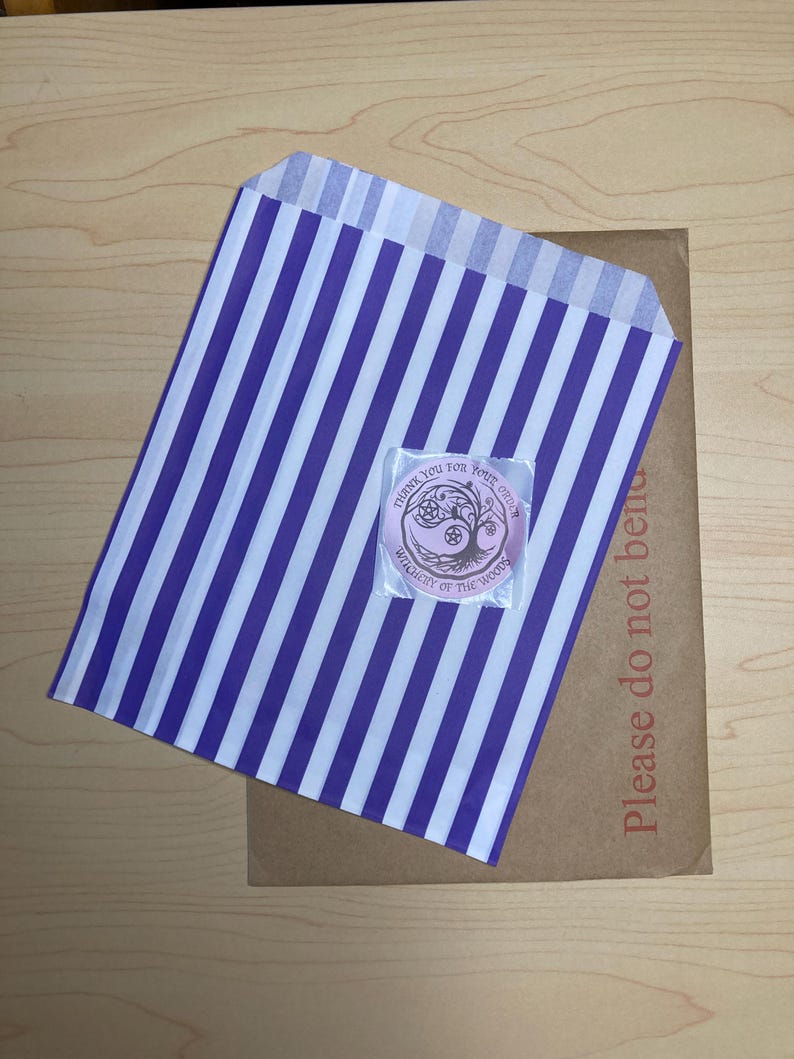 May include: A purple and white striped paper bag with a small sticker on top. The sticker has text that reads "Thank you for your order" and "Witchery of the Woods". A brown cardboard mailer is underneath the bag, with the text "Please do not bend".