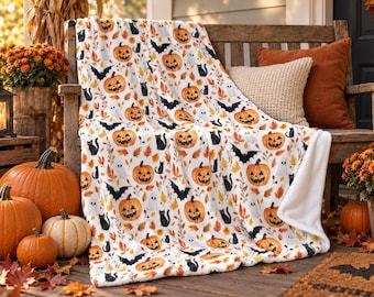 Op de afbeelding: Een witte plaid met een herhalend patroon van oranje pompoenen, zwarte katten, zwarte vleermuizen, witte spoken en kleurrijke herfstbladeren. De deken heeft een zachte textuur en is gedrapeerd over een houten bank.