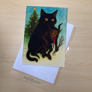 May include: Greeting card featuring a black cat holding a Krampus figure with a long red tongue. A small Christmas tree is visible on the left. The card is on top of a white envelope and a clear plastic sleeve.