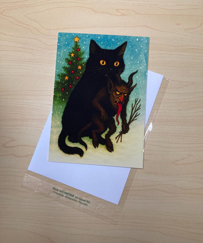 May include: Greeting card featuring a black cat holding a Krampus figure with a long red tongue. A small Christmas tree is visible on the left. The card is on top of a white envelope and a clear plastic sleeve.