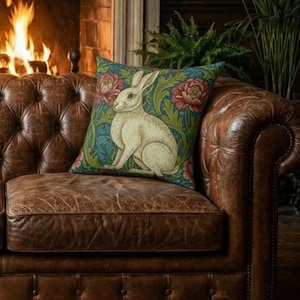 Puede incluir: Cojín cuadrado decorativo con un conejo crema sobre un fondo verde azulado con detalles florales rojos y verdes. El cojín está colocado sobre un sofá Chesterfield de cuero marrón, creando una estética vintage y acogedora.