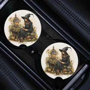 May include: Two round car coasters featuring a witch in a black dress and pointed hat reading a book in a forest setting. The coasters are white with a brown border and the text "Witchcraft of the Woods" is printed on the bottom.