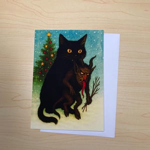 May include: A greeting card featuring a black cat holding a Krampus-like creature with a long red tongue. A Christmas tree with a star and red ornaments is in the background. The card is on a wooden surface.