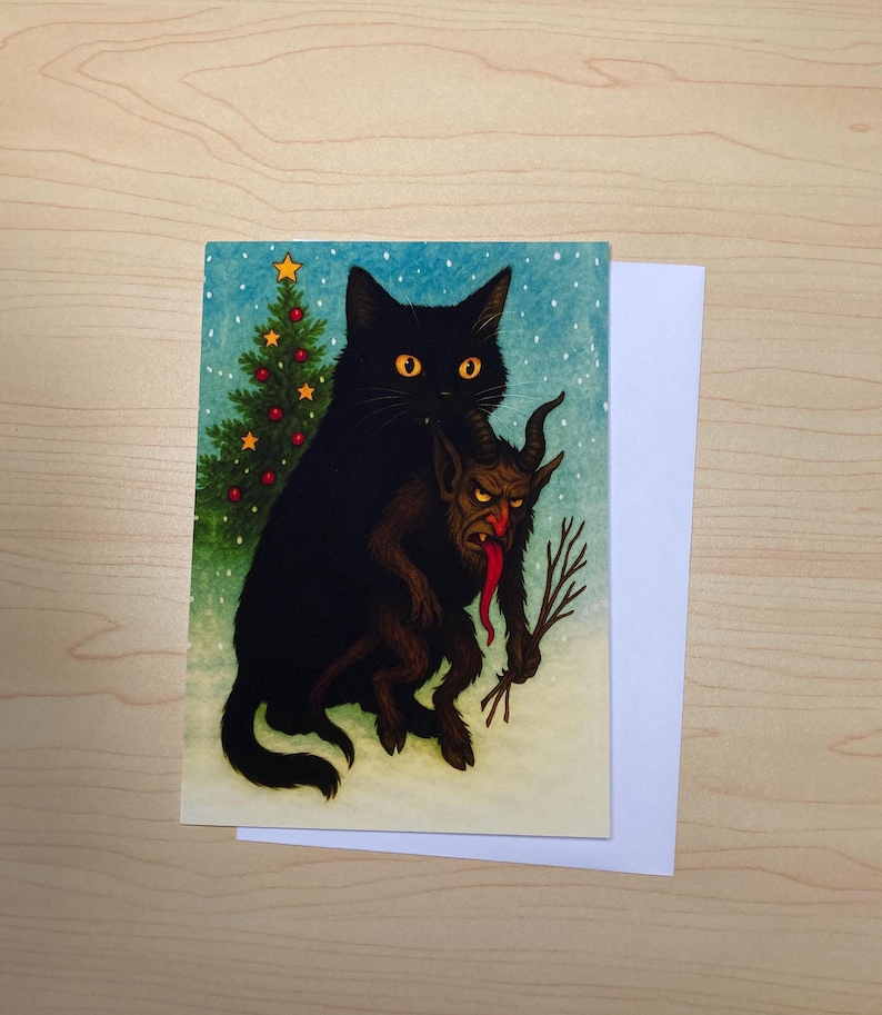 May include: A greeting card featuring a black cat holding a Krampus-like creature with a long red tongue. A Christmas tree with a star and red ornaments is in the background. The card is on a wooden surface.