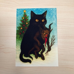 Op de afbeelding: Een grillige illustratie toont een zwarte kat die een Krampus-achtig figuur vasthoudt. Een kleine kerstboom met rode ornamenten en een ster staat links. De achtergrond is een besneeuwde scène met een blauwe lucht.