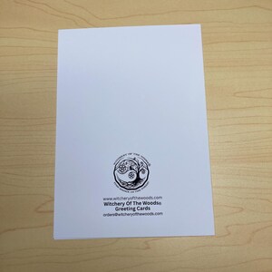 May include: A white greeting card with the text "Witchery Of The Woods&reg; Greeting Cards" and a circular logo featuring a tree and a pentacle. The card is on a light brown wooden surface.