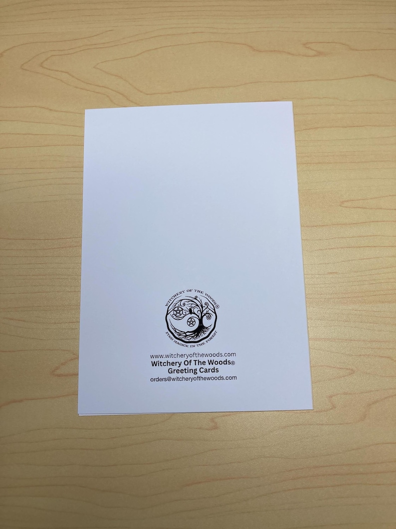 May include: A white greeting card with the text "Witchery Of The Woods&reg; Greeting Cards" and a circular logo featuring a tree and a pentacle. The card is on a light brown wooden surface.