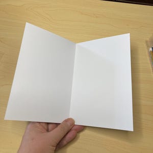 May include: A blank, white greeting card, open to reveal the inside. The card is held open, displaying the clean, unfolded pages. The card is on a light wood surface.