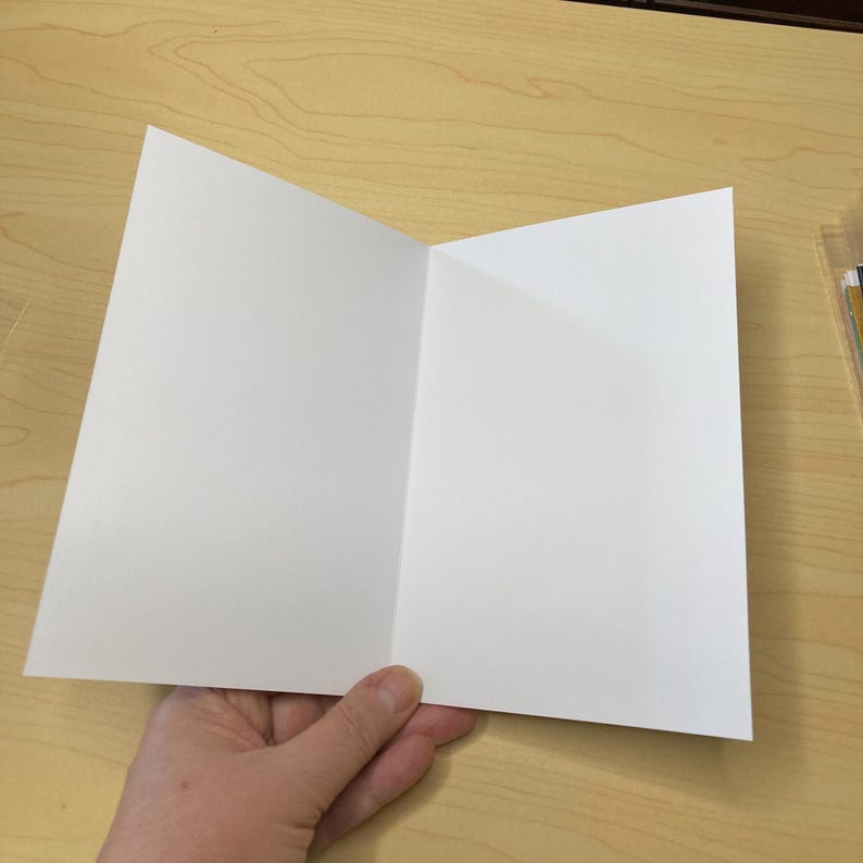 May include: A blank, white greeting card, open to reveal the inside. The card is held open, displaying the clean, unfolded pages. The card is on a light wood surface.