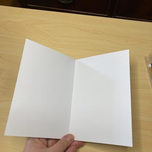 May include: A blank, white greeting card, folded in half, held by a hand. The card is open, displaying two blank panels. The background is a light wood surface with a dark brown cabinet in the background.