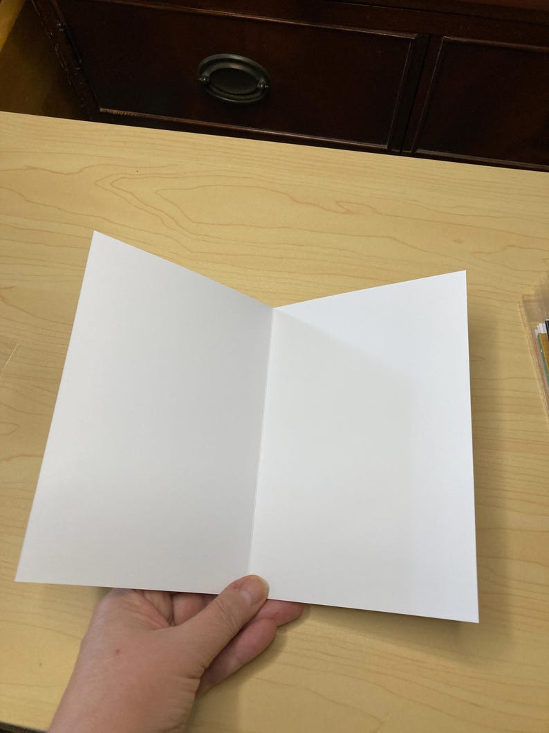 May include: A blank, white greeting card, folded in half, held by a hand. The card is open, displaying two blank panels. The background is a light wood surface with a dark brown cabinet in the background.
