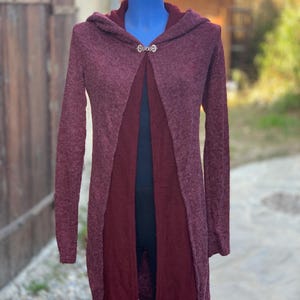 Op de afbeelding: Een lange, bordeauxrode cardigan met een capuchon en een zilveren sluiting. De cardigan is open aan de voorkant en heeft een iets langere rug dan de voorkant.