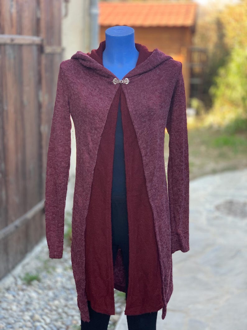 Op de afbeelding: Een lange, bordeauxrode cardigan met een capuchon en een zilveren sluiting. De cardigan is open aan de voorkant en heeft een iets langere rug dan de voorkant.